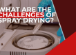 A person wearing blue gloves and a lab coat is seen adding white powder to a sample pan inside a laboratory instrument, with overlaid text asking: 'WHAT ARE THE CHALLENGES OF SPRAY DRYING?'