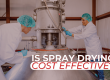 Two scientists in lab coats and hairnets working on a large industrial spray dryer. Overlaid white and red text asks: 'IS SPRAY DRYING COST EFFECTIVE?'